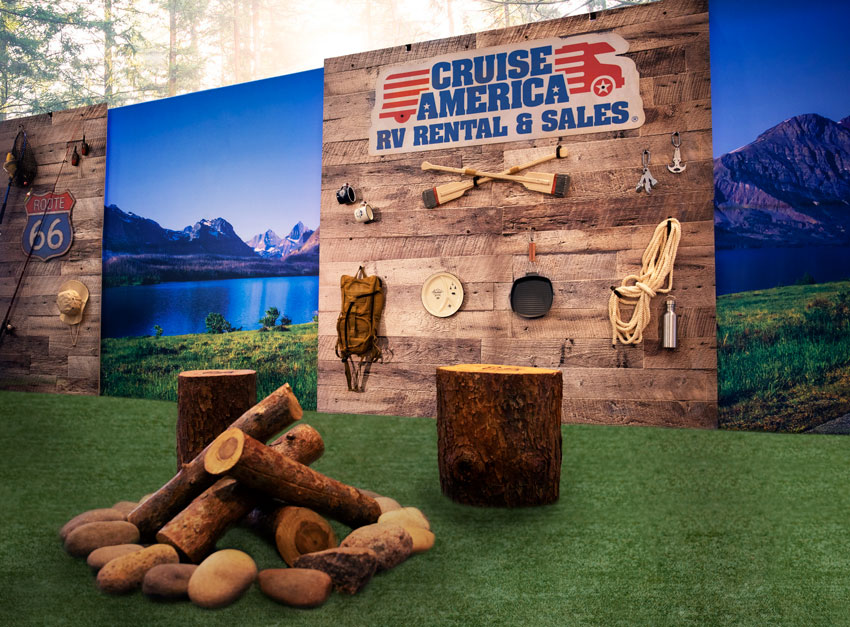 A tradeshow booth with camping gear attached to wood walls. A campfire and tree stump sit on grass in font of the booth.